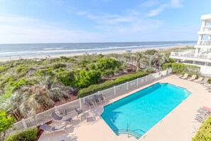 A pool area with a view of the beach and ocean, surrounded by greenery and a white fence. - Oceanfront Condo just steps to beach, pool, and main strip - Ocean Palms 201 (Isle of Palms)