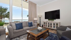 A living room with a sofa, coffee table, and TV. There is a balcony with a view of palm trees. - Lobby-level, coastal contemporary beachfront condo! 🏖️ (Destin)