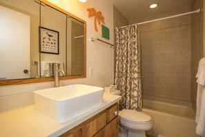 A bathroom with a white sink, a toilet, a shower with a patterned curtain, and a mirror with a framed picture of marine life.