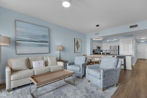 A modern living room with a sofa, armchairs, and a coffee table. A kitchen is visible in the background with white cabinets and a dining area. - Gulf Views-Short Walk 2 Beach-Resort Amenities (Miramar Beach)