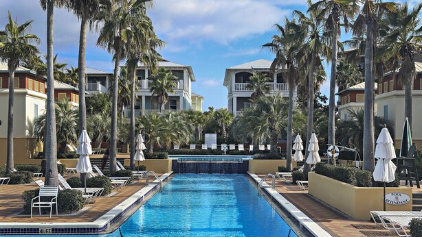 Ein Swimmingpool mit Liegestühlen und Sonnenschirmen, umgeben von Palmen und Hotelgebäuden.