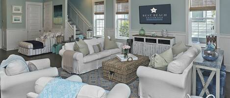 A spacious living room with a white sofa set, a coffee table, and a wicker basket. There is a staircase in the background leading to another level.
