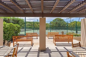 Ein Sitzbereich im Freien mit Holzbänken und -tischen, einer Pergola darüber und ein Tennisplatz, der durch einen Zaun sichtbar ist.