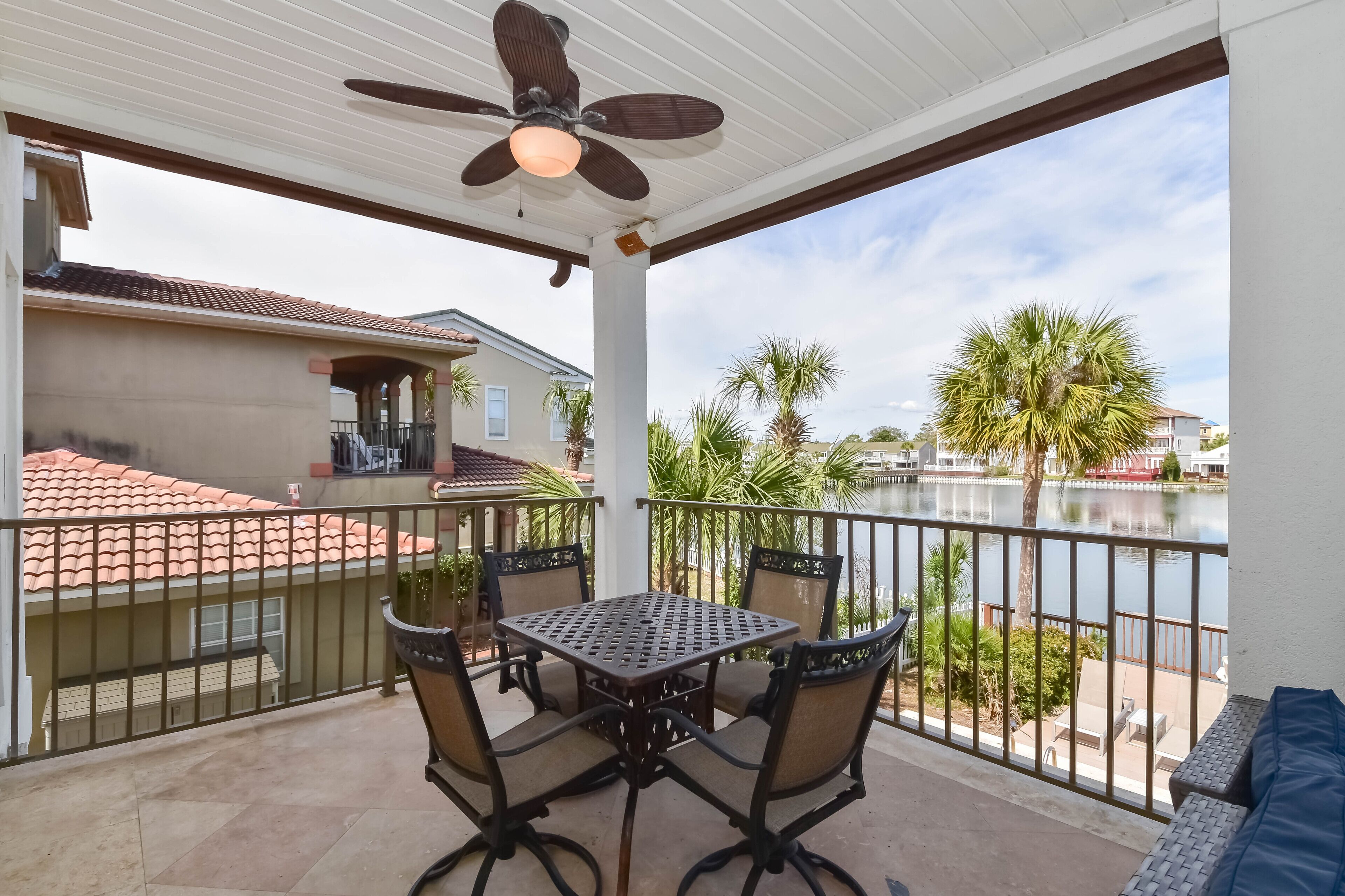 A covered balcony with a table and chairs, a ceiling fan, and a view of a lake and palm trees.