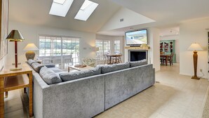 Ein geräumiges Wohnzimmer mit einem großen, grauen Ecksofa, einem Kamin und einem darüber montierten Fernseher. Zwei Dachfenster sorgen für natürliches Licht.