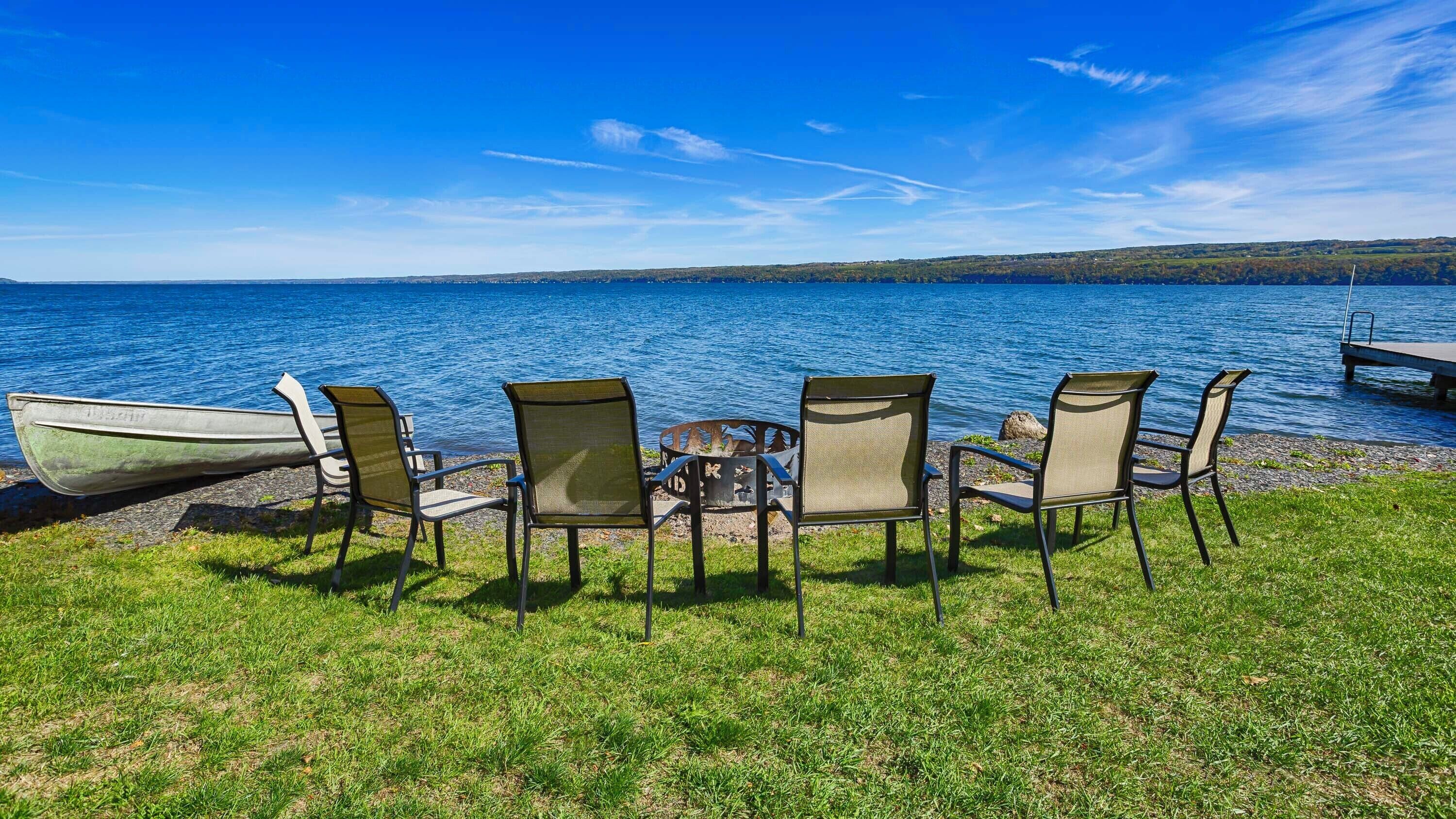 Een zitgedeelte aan het meer met vier stoelen en een kleine vuurkuil, met uitzicht op een rustig meer met een steiger op de achtergrond.