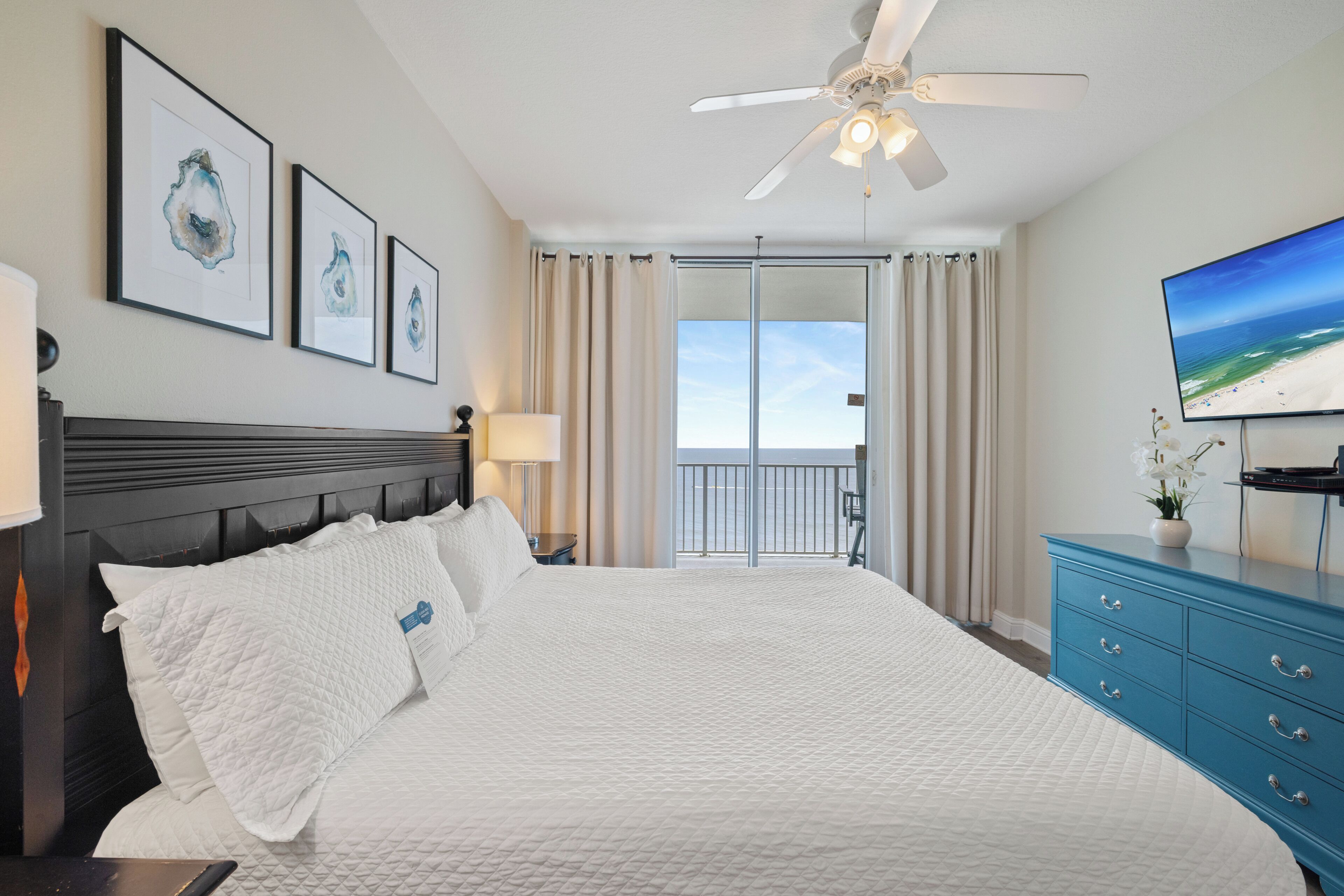 A hotel room with a large bed, a ceiling fan, a TV, and a view of the beach.