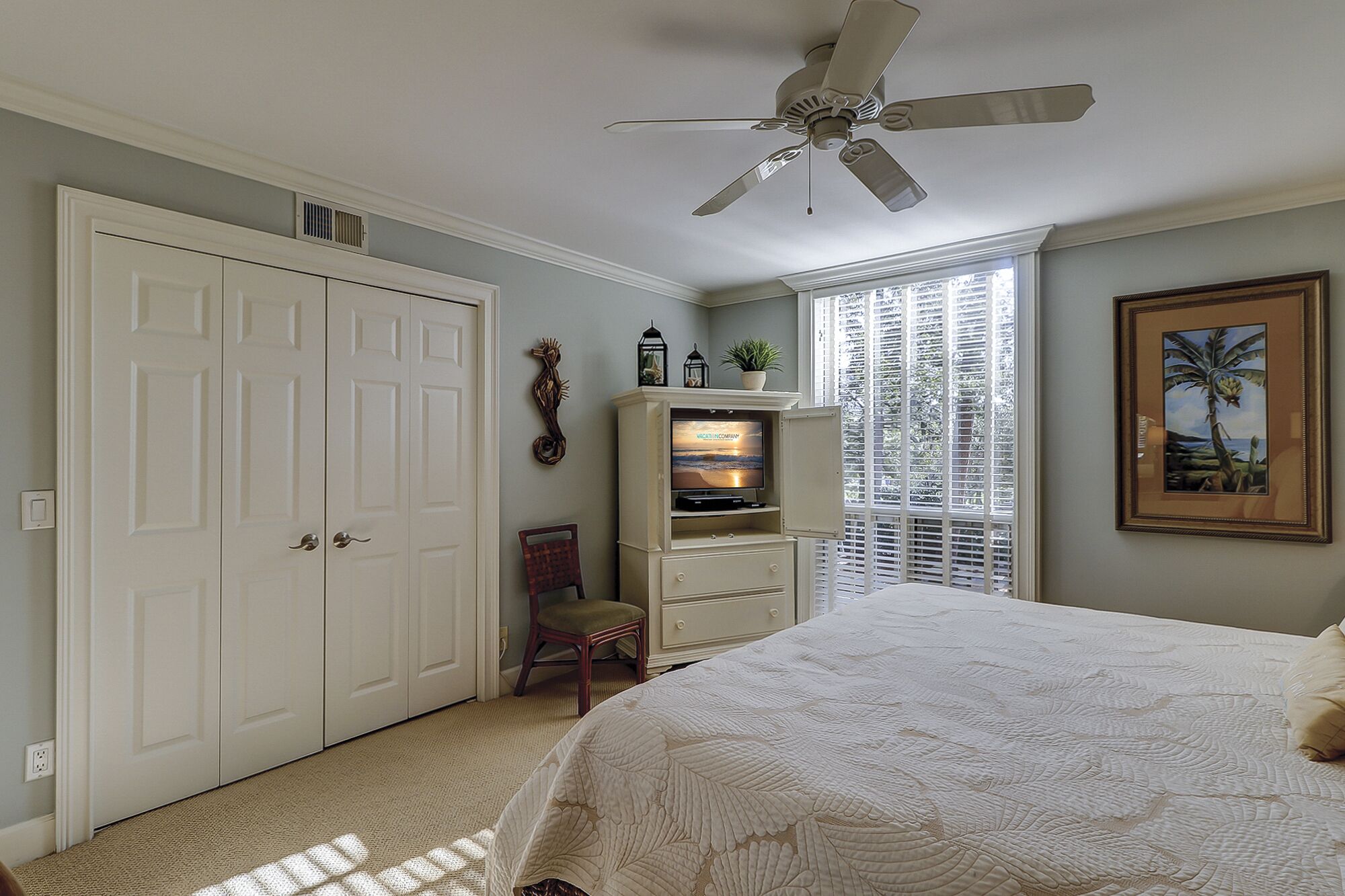 Un dormitorio con una cama, ventilador de techo, televisión en mueble, una silla y ventana con persianas.