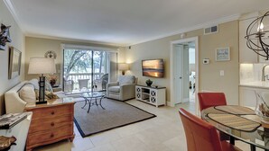A spacious living room with a large window, a dining area with a glass table, and a kitchen area. - 913 Cutter Ct- Short Stays All Summer - Steps to Harbour Town and Marina (Hilton Head Island)