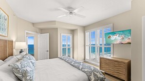 A bedroom with a bed, a ceiling fan, a TV, and a view of the ocean through large windows. - Wraparound Balcony ~ Condo Remodel Jan-Feb 2026 (Miramar Beach)