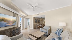 A spacious living room with a flat-screen TV, a ceiling fan, a dining area, and a kitchen in the background. - Wraparound Balcony ~ Condo Remodel Jan-Feb 2026 (Miramar Beach)