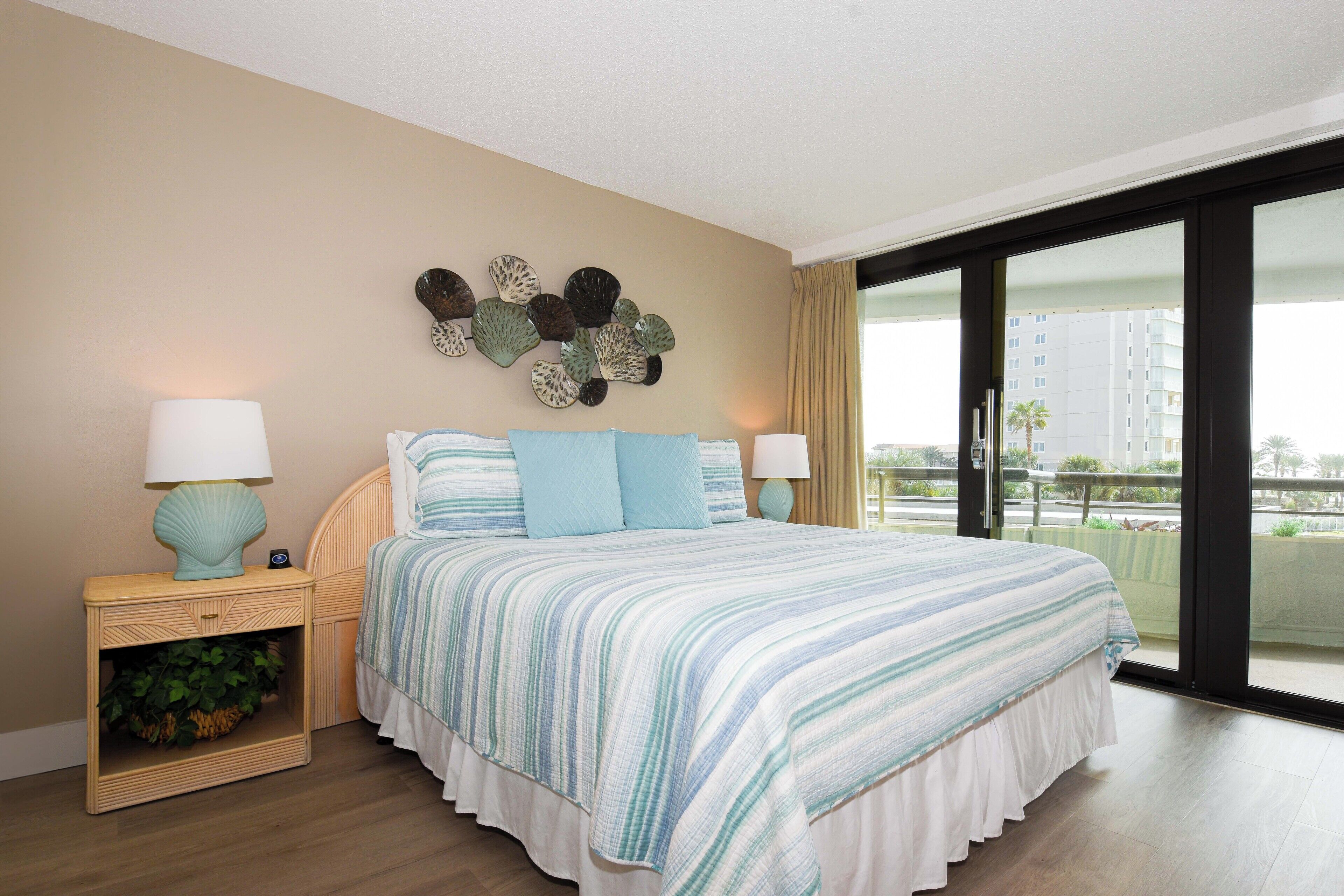 Una habitación de hotel con una cama grande, dos mesitas de noche con lámparas, un adorno en la pared y vistas a un edificio a través de un ventanal amplio.