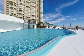 Una piscina de borde curvo, una zona junto a la piscina en blanco y un edificio alto al fondo.