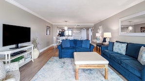 A living room with a TV, blue sofas, a coffee table, and a kitchen in the background.