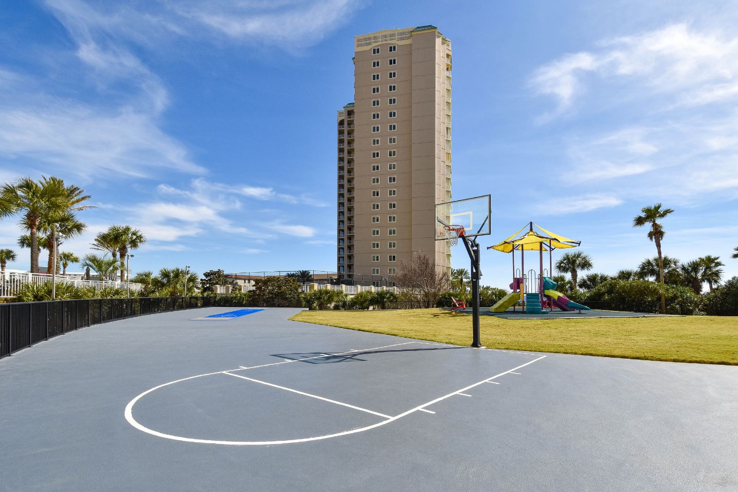 A basketball court with a hoop, a playground with a slide, and a tall building in the background.