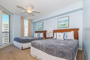 Quarto de hotel com duas camas, um ventilador de teto, janelas amplas e obras de arte nas paredes.