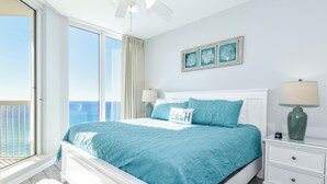 A bedroom with a large bed, a nightstand, a lamp, and a view of the ocean through a sliding door.
