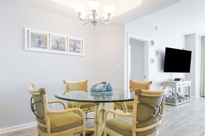 Uma sala de jantar com mesa de vidro redonda e cadeiras de vime, um candelabro, uma televisão de tela plana e uma pequena mesa branca com gavetas.
