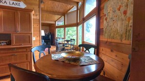 Interieur van een houten hutje met een eettafel, stoelen, een groot raam en een bord waarop 'KA LAKE' staat.