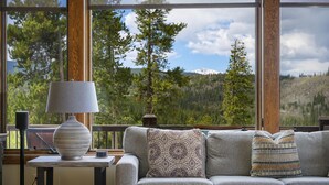 A room with a sofa, a table with a lamp, and a view of a mountainous landscape through large windows.