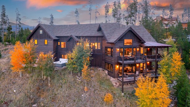 A large, multi-story wooden cabin with a balcony, surrounded by trees and a mountainous backdrop.