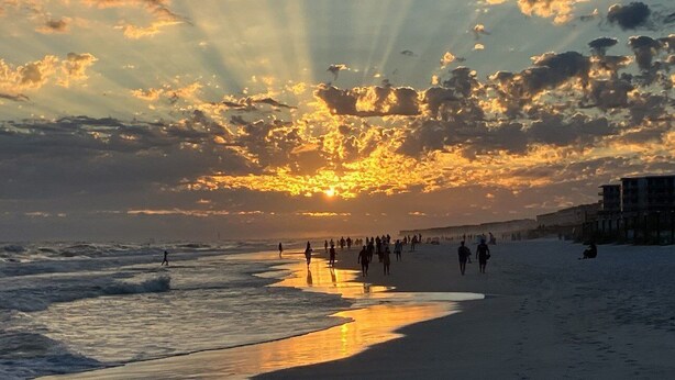 Uma praia ao pôr do sol, com ondas e pessoas caminhando pela orla.