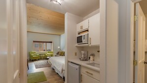 Un dormitorio compacto con cama, mesita de noche y ventana. Junto al dormitorio, una kitchenette con microondas, cafetera y un refrigerador pequeño.
