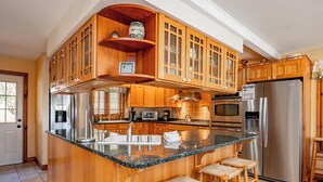 A kitchen with wooden cabinets, a stainless steel refrigerator, a granite countertop, and bar stools. - Dog Friendly Lakefront Home w/ Dock & Hot Tub (MCHENRY)
