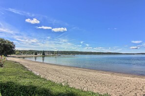 A sandy beach with a calm lake, a grassy area with trees, and a distant shoreline with buildings and wind turbines.