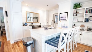 A modern kitchen with white cabinetry, a central island with bar stools, a stainless steel trash can, and a wooden floor.