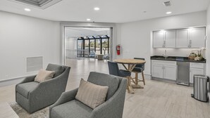 Una sala de estar moderna con sofás grises, una mesa de comedor de madera y una zona de cocina con armarios blancos y un lavavajillas de acero inoxidable.