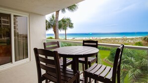 Un balcón con mesa y sillas de madera, con vistas a una playa con palmeras y al océano.