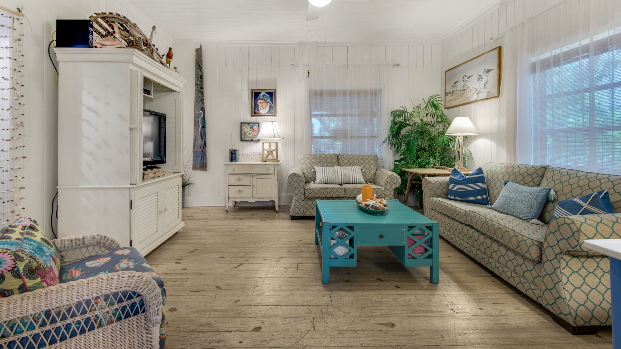 A cozy living room with a television, a patterned sofa, a teal coffee table, and a wicker chair.
