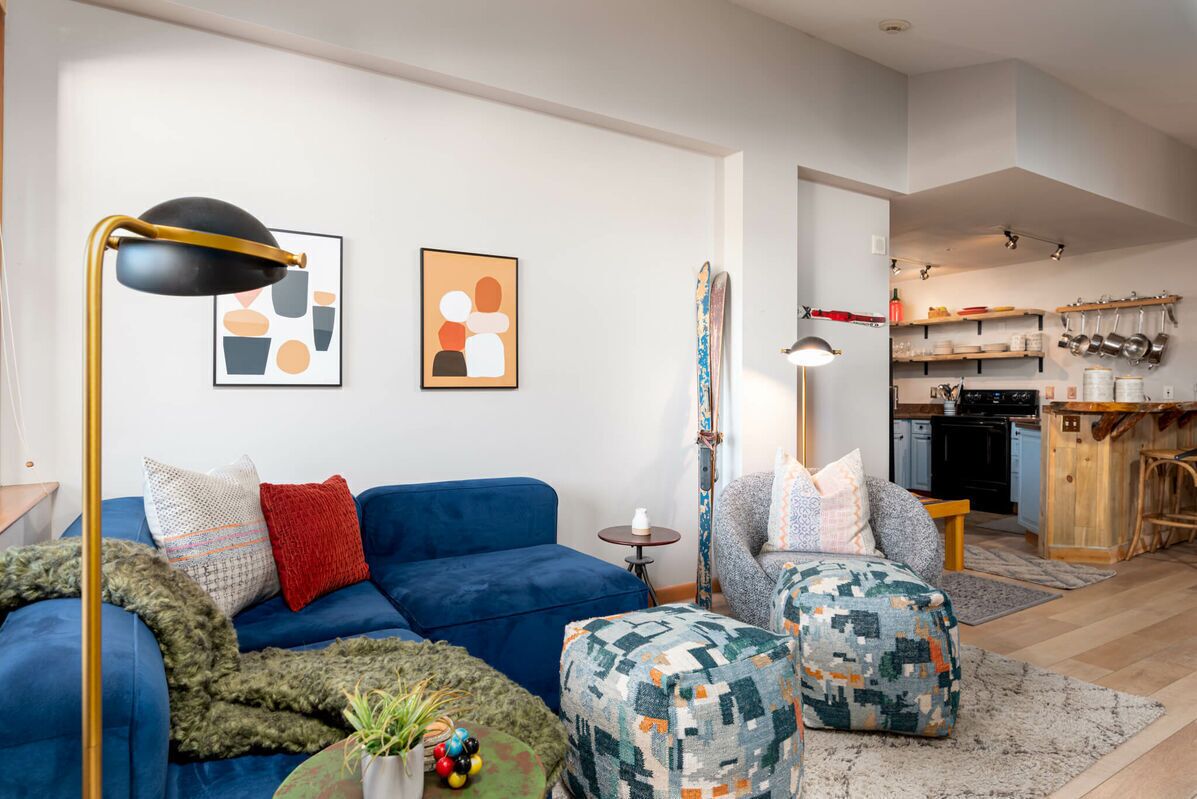 A modern living room with a blue sofa, patterned ottomans, a floor lamp, and abstract wall art.