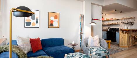 A modern living room with a blue sofa, patterned ottomans, a floor lamp, and abstract wall art.
