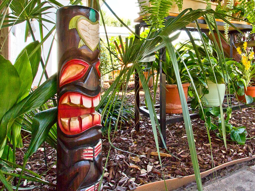 Una escultura de madera con rostro tallado, rodeada de plantas en macetas y follaje.