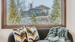 A room with a window overlooking a modern house, a gray sofa with two patterned throw pillows, and a green blanket.