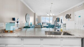 Una cocina moderna con encimera de mármol, armarios blancos y una isla central. Hay un comedor con sillas azules y vistas al exterior.