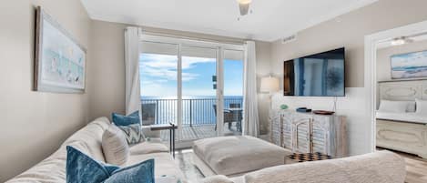 A spacious living room with a large window overlooking the ocean, a ceiling fan, and a television on the wall.