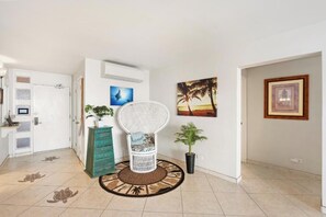 A hallway with a white door, a white chair with a cushion, a green chest of drawers, a round rug, and two potted plants.