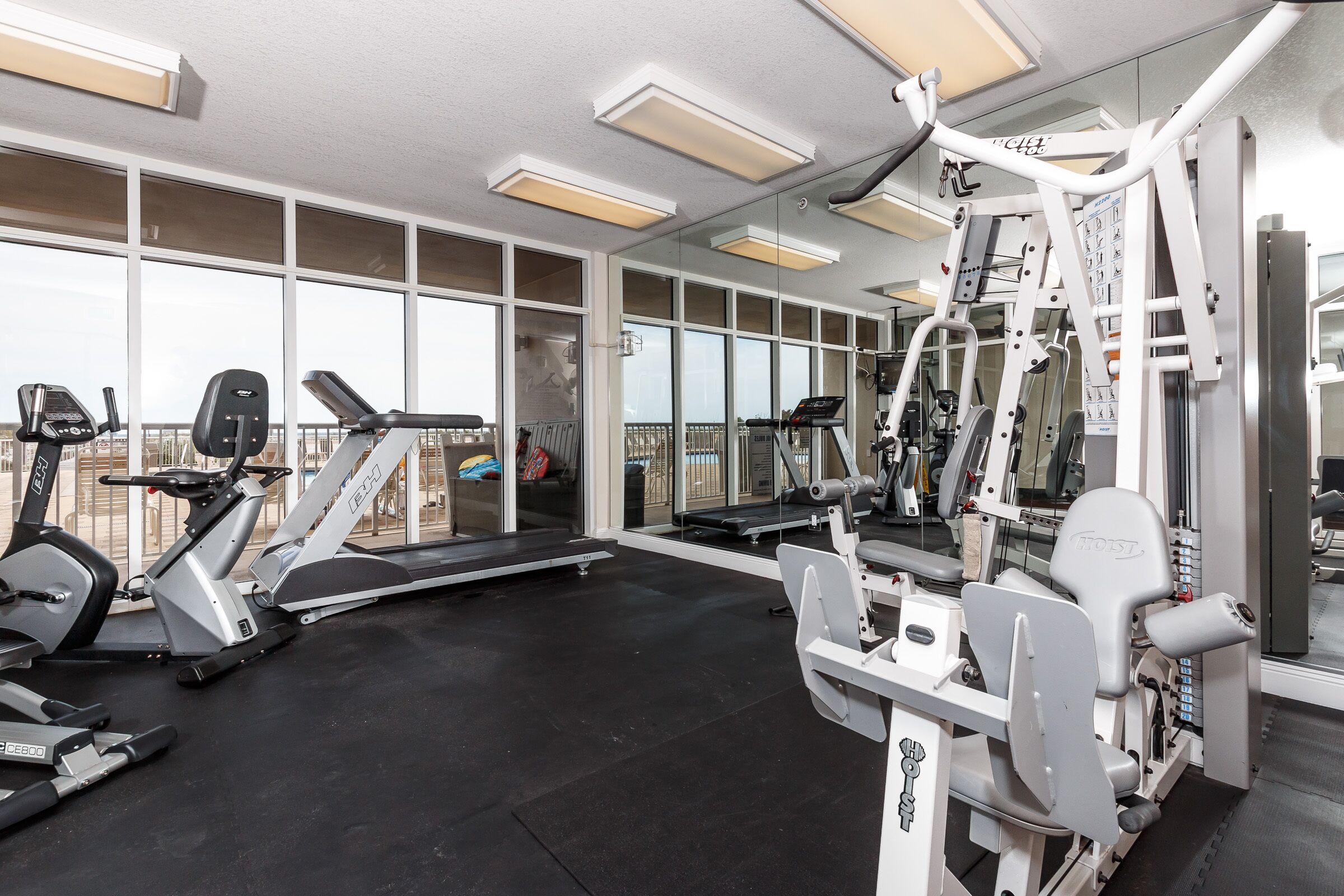A gym with various exercise machines, large windows, and a mirrored wall.