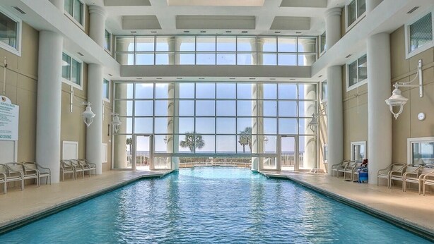 Piscina interna com vista clara para o oceano através de grandes janelas, rodeada por cadeiras de descanso.