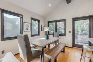 A modern dining area with a wooden table, chairs, and a bench. There are large windows providing natural light and views of the outside.