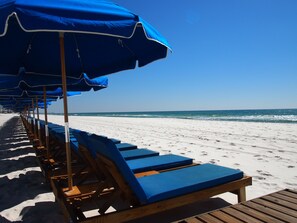 Una playa con filas de sillas azules y sombrillas, una terraza de madera y una vista despejada del océano.