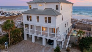 Une maison de deux étages située en bord de mer, dotée d’un balcon et d’une véranda couverte, et se trouvant à proximité de la plage, où des véhicules sont stationnés.