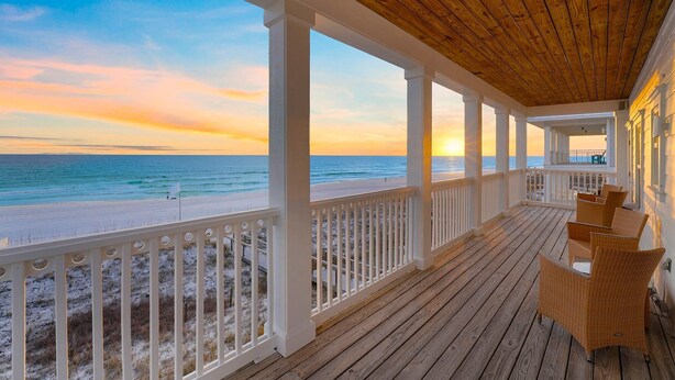 Une véranda couverte avec un plafond en bois et des balustrades blanches, donnant sur une plage et offrant une vue sur le coucher de soleil.