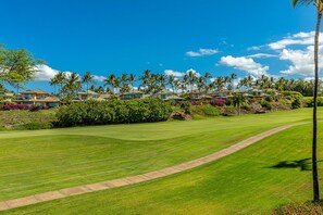 Um campo de golfe com um green bem cuidado, céu claro e uma fileira de villas ao fundo.