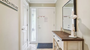 A hallway with a sign reading "BEACH HOUSE," a white door with a "No Smoking" sign, a wooden vanity with a mirror, and a blue doormat.