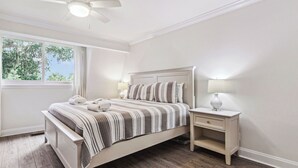 A bedroom with a large bed, a nightstand, a ceiling fan, and a window with a view of trees.
