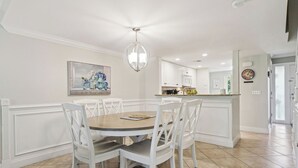 A dining area with a round table, white chairs, a chandelier, and a kitchen in the background.